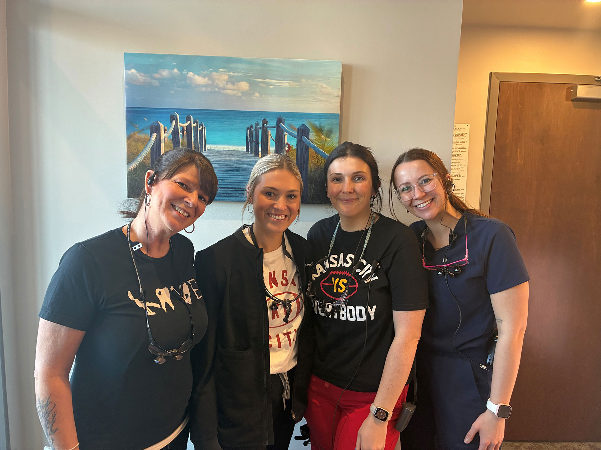 A group of four people posing together with smiles, standing in front of a painting, with one person wearing a stethoscope and another holding a camera.