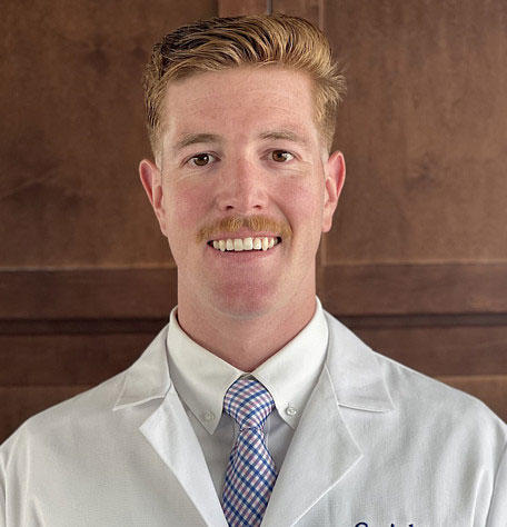 The image shows a man with a mustache wearing a white lab coat and a tie, smiling at the camera.