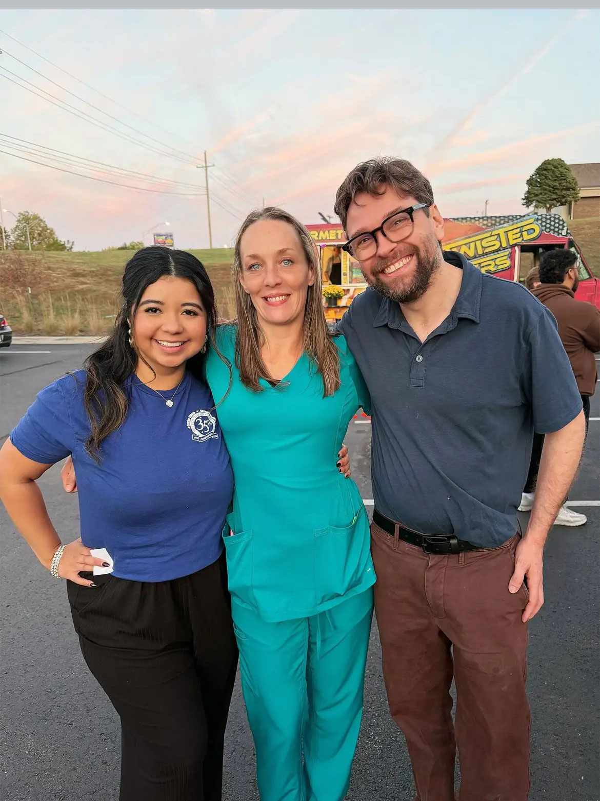 The image shows three individuals standing together outdoors during sunset two adults appear to be posing for a photo, one wearing a blue shirt and the other in scrubs with a stethoscope around their neck, while a third person stands behind them, smiling and wearing a white shirt with a logo.
