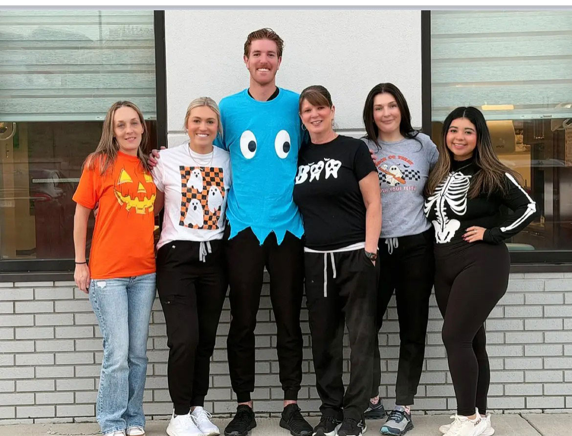 The image features a group of people posing together, with some individuals wearing costumes, including one person dressed as Pac-Man, standing in front of a storefront with a Halloween theme.