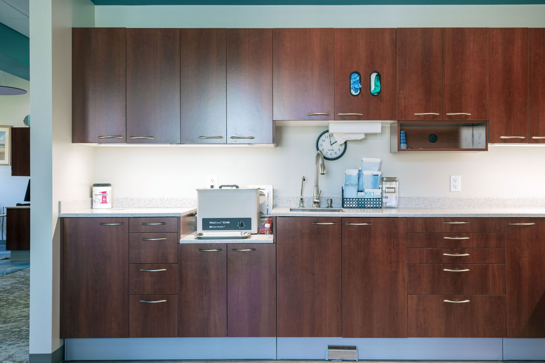 The image shows a modern kitchen with wooden cabinets, a countertop with a sink, and various appliances such as a microwave and coffee maker.