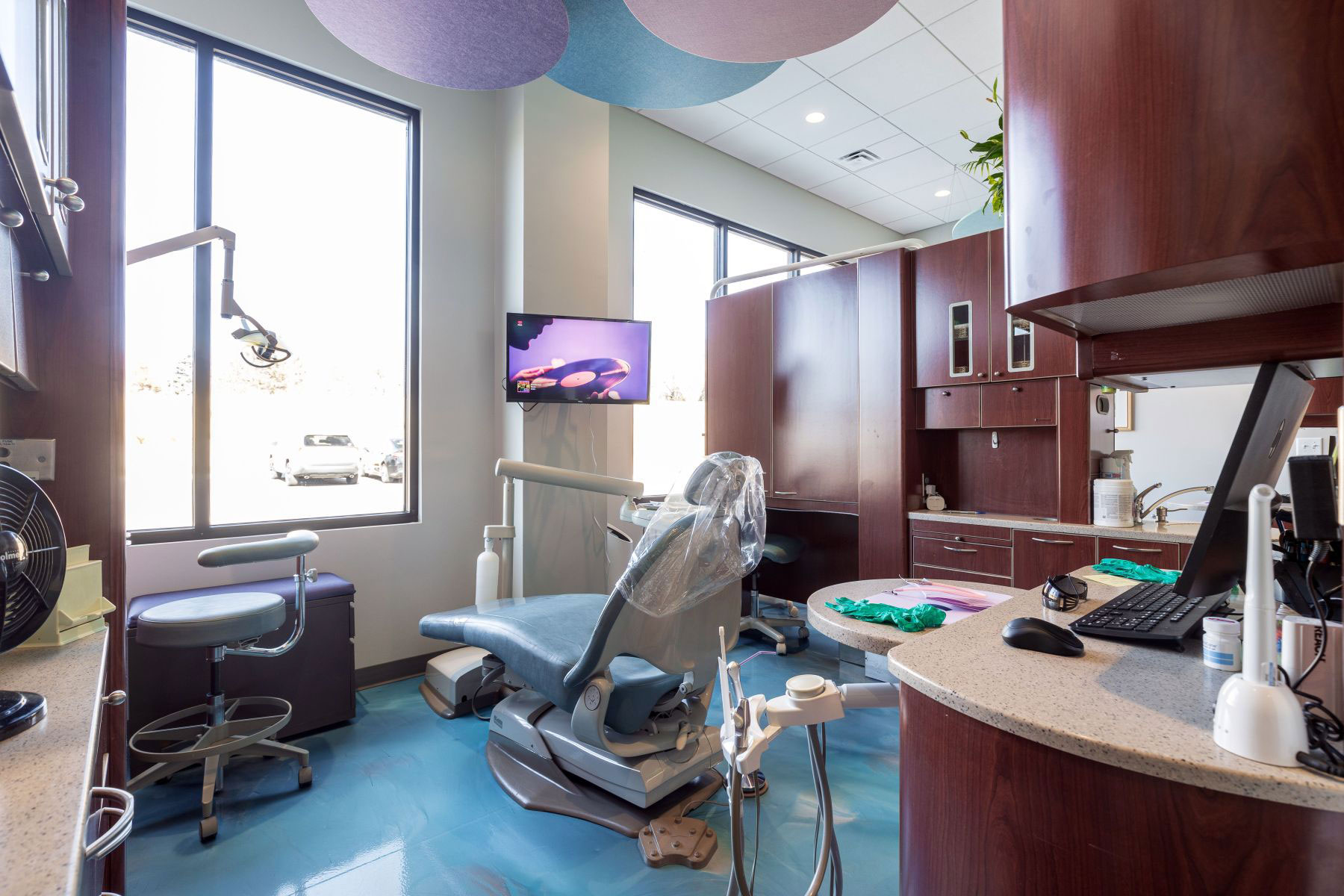 The image depicts an interior view of a dental office with modern furnishings and equipment, featuring a reception area, dental chairs, and a window with natural light.